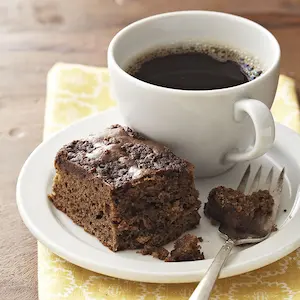 Weißer Teller mit einem Stück Schokoladenkuchen und einer Gabel, daneben eine weiße Tasse mit schwarzem Kaffee.
