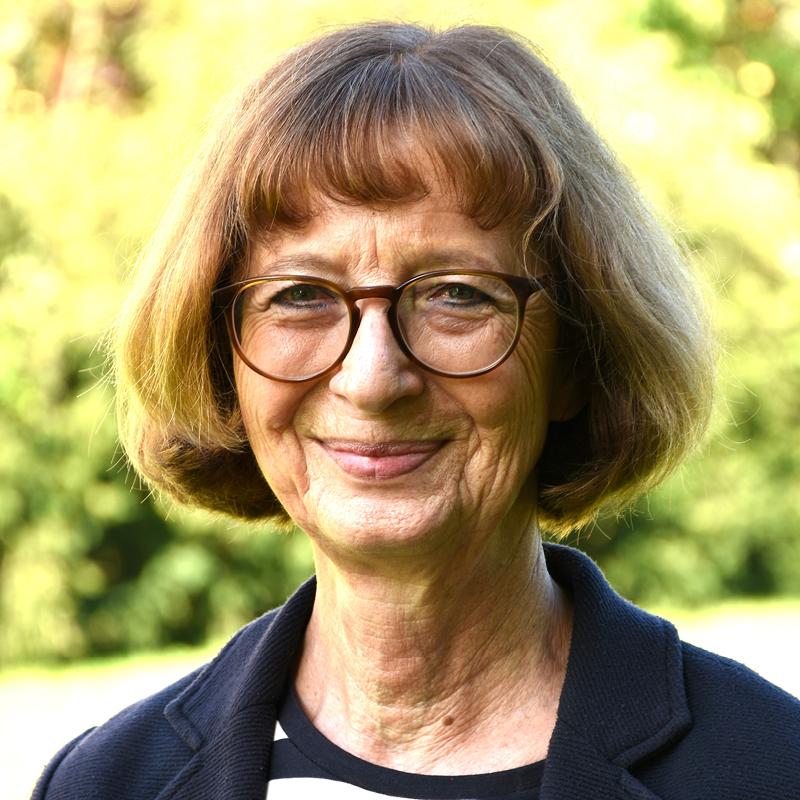 Christine Barth Porträt einer älteren Frau mit kurzen braunen Haaren, Brille und gestreiftem Oberteil vor unscharf grünem Hintergrund.