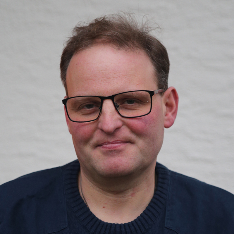 Peter Hertnagel Porträt eines Mannes mit kurzen braunen Haaren, Brille und dunkelblauem Pullover vor einem hellen Hintergrund.
