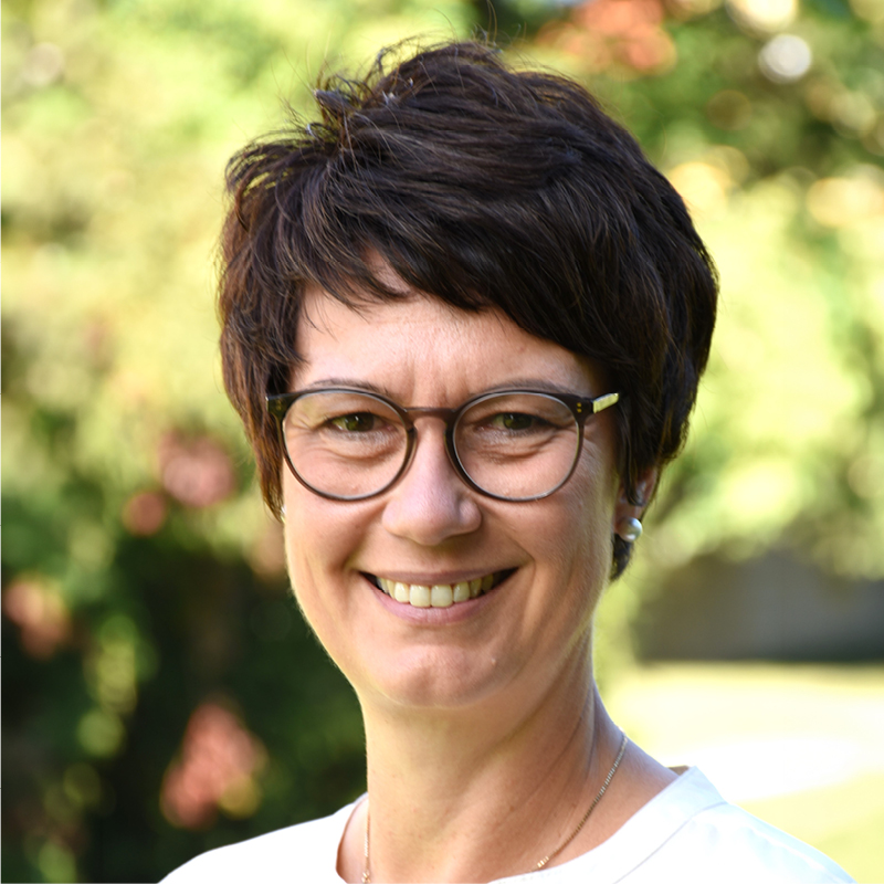 Karin Winkler Porträt einer lächelnden Frau mit kurzen dunklen Haaren und Brille vor unscharf grünem Hintergrund im Freien.